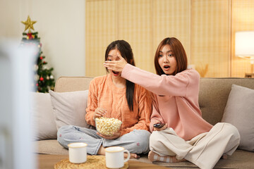 Young woman lesbian couple in sweater sits on comfortable sofa to covering eyes girlfriend and watching