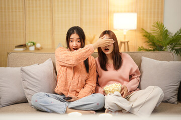 Young woman lesbian couple in sweater sits on comfortable sofa to covering eyes girlfriend and watching