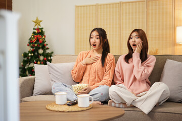 Young woman lesbian couple in sweater sitting on comfortable sofa watching scary horror cinema with