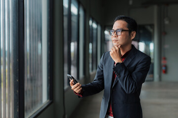Asian businessman in glasses thinking deeply, holding a smartphone in an office corridor, contemplating business decisions