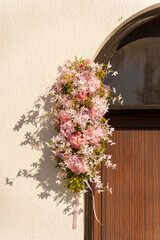Soft pink flowers create a lovely arrangement hanging on a wooden door. The sunlight highlights the delicate blooms and adds charm to the entrance of the building.
