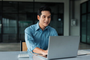 Asian businessman typing on laptop, smiling, working remotely or in a modern startup office, showcasing professionalism and technology