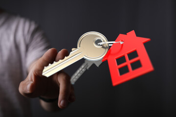A person's hand presenting keys with a red house tag symbolizing real estate, home ownership, and the concept of buying and renting a house.