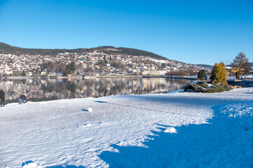 Obraz premium serene winter lakeside scene with snow-covered banks, blue water lake gerardmer, French Vosges, forested hills in distance, frozen landscape snow, still water, and bare trees, Seasonal beauty france