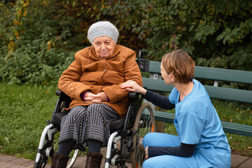 Elderly woman in wheelchair with healthcare worker in blue uniform outside in park