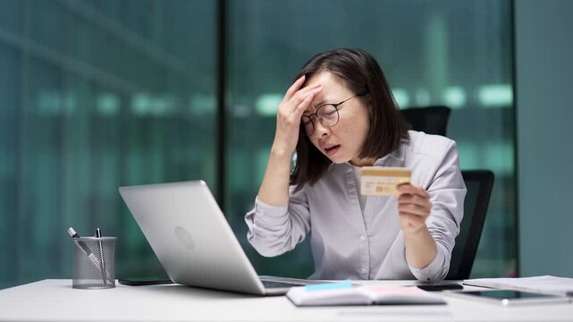 Shocked worried Asian businesswoman detected fraud checking credit card on laptop at workplace in business office. Upset female employee became a victim of deception, money was stolen from her account