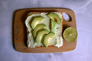 square table of bread with spread avocado butter with avocado slices on wooden board on dark background