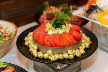 Colorful layers of sliced tomatoes and cheese create an appealing salad centerpiece for a festive gathering, surrounded by fresh herbs and other dishes.