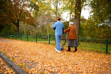 Obraz premium Elderly woman taking a walk through the park in autumn with help of walker and caretaker aid