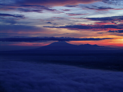 Aerial view of the majestic silhouette of Lawu Mountain piercing through a sea of ethereal clouds under a vibrant sunrise sky, Karanganyar Regency, Central Java, Indonesia.