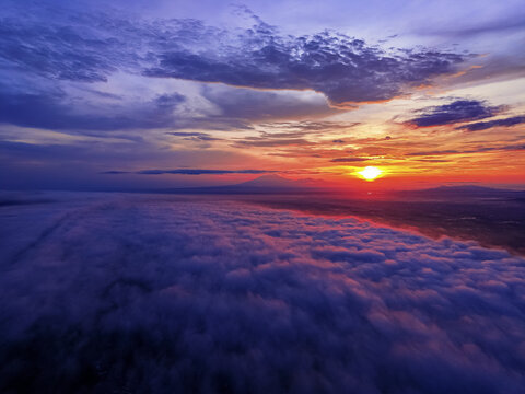 Aerial view of a breathtaking sunrise over a thick blanket of clouds, with the sun's radiant glow contrasting against the distant silhouette of Lawu Mountain, Karanganyar, Central Java, Indonesia.