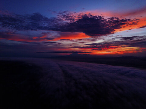 Aerial view of the lawu mountain peak piercing through the thick blanket of clouds under a fiery sunset sky, Karanganyar Regency, Central Java, Indonesia.
