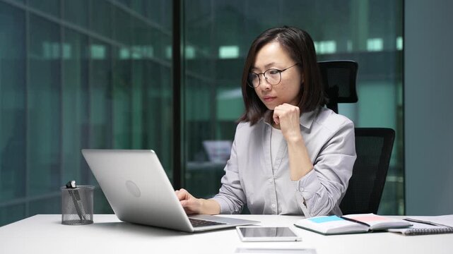 Thoughtful asian businesswoman thinking about problem solving while working on laptop computer sitting at desk at workplace in a modern business office. Female manager is engaged in project or startup