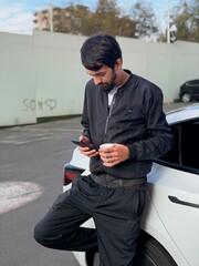 A young man is leaning against the back fender of a white car in an outdoor urban setting. He is looking intently at his mobile phone, holding a disposable coffee cup in his other hand. 