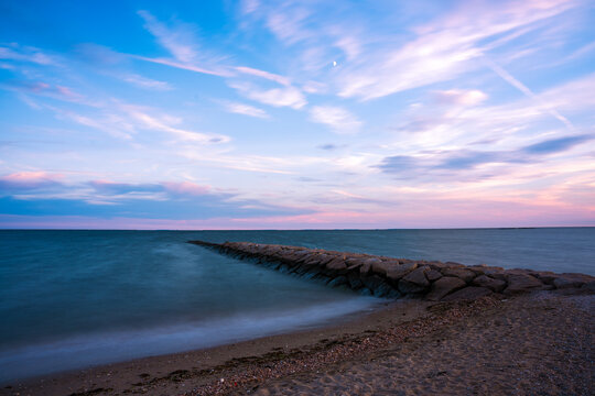 View of a stone jetty stretches into the tranquil ocean under a pastel sky brushed with pink and blue hues, captured from the sandy shore, Madison, Connecticut, United States.
