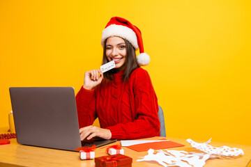 Playful christmas shopper girl with santa hat smiling at laptop while gifting decor on a bright...