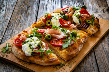 Pinsa with cured ham, burrata cheese, tomatoes, green olives and arugula on wooden table	