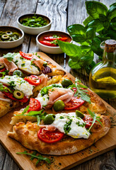 Pinsa with cured ham, burrata cheese, tomatoes, green olives and arugula on wooden table	