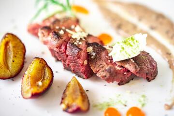 Medium-rare steak, sliced, with dogwood sauce on a white plate with roasted plums and rosemary cream cheese dumplings. Top view.