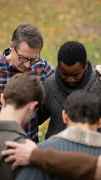 Group of diverse men praying together outdoors in unity, heads bowed and arms around shoulders, symbol of faith, brotherhood, and spiritual support in community