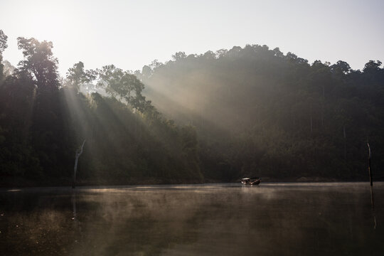 View of sun rays piercing through the misty air over tranquil waters, reflecting the silhouette of a boat against the lush, green hillsides, Surat Thani, Surat Thani, Thailand.