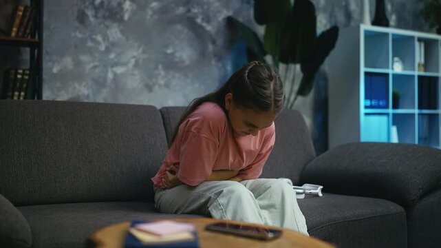 Sad teenage girl suffering lower abdominal pain while sitting on couch at home