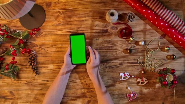 Top view of hands swiping a smartphone with a green screen on a rustic wooden table surrounded by Christmas decorations and wrapping materials, ideal for festive shopping or design layouts.