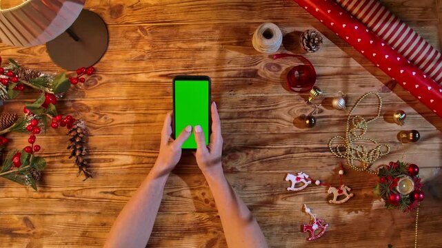 Top view of hands typing a smartphone with a green screen on a rustic wooden table surrounded by Christmas decorations and wrapping materials, ideal for festive shopping or design layouts.