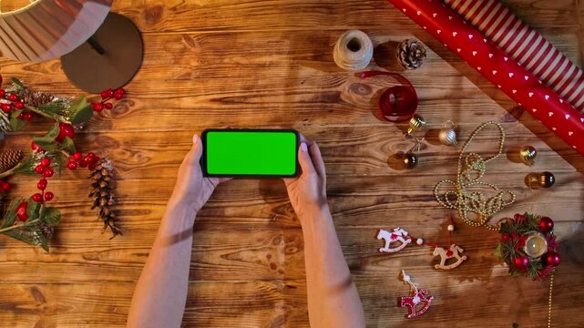 Top view of hands holding a smartphone with a green screen on a rustic wooden table surrounded by Christmas decorations and wrapping materials, ideal for festive shopping or design layouts.