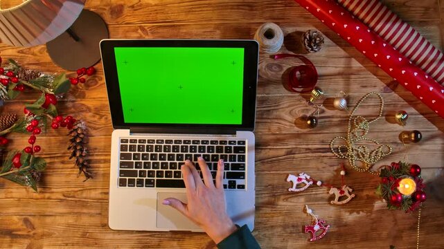 Top view of hands works on a laptop with a chromatic green screen on a wooden table surrounded by packaging materials and decorations. The perfect layout for holiday shopping or design.