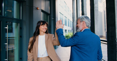 Two colleagues of adult man and young woman, walk toward each other outside a modern office building, smiling cheerfully, exchanging a high five, then parting ways to continue their day