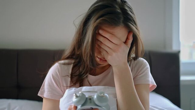 Tired young woman sitting on bed holding an alarm clock early in the morning, feeling sleepy and exhausted after waking up.