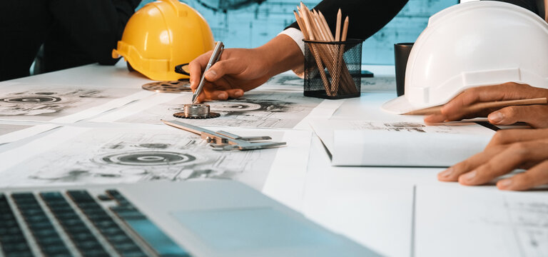 A focused team of engineers collaborates on detailed construction plans in an office environment, using safety helmets and modern technology for effective project execution. SACTR - Powered by Adobe