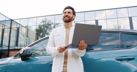 Caucasian man working remotely on laptop while standing next to his parked car outside modern office building, appearing focused and engaged in his task, exemplifying the flexibility of mobile work