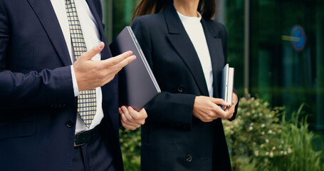 Close-up of two business partners, a man and a woman in formal suits, walking together in an urban area while holding digital devices, outdoor