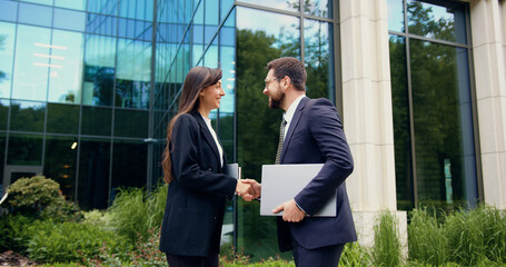 Close-up of two business partners, a man and a woman in formal suits, greeting each other with a handshake outdoors near an office building in the city.