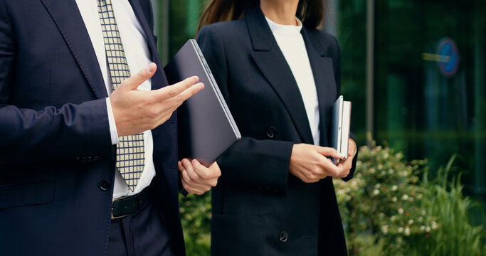 Close-up of two business partners, a man and a woman in formal suits, walking together in an urban area while holding digital devices, outdoor - Powered by Adobe