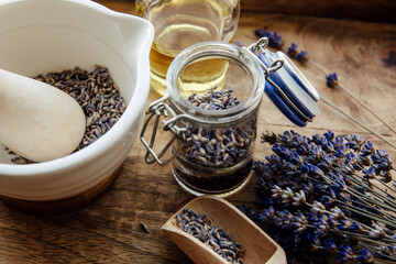Dried lavender flower buds inside clear glass jar, with mortar and pestle filled with flower and bouquet next to it. Brown wood background.