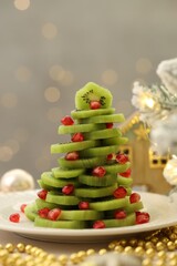 Christmas tree made of kiwi, pomegranate seeds and festive decor on table against light background with blurred lights, closeup. Bokeh effect