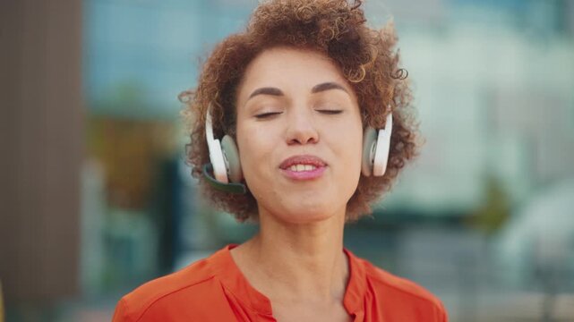 Smiling African American woman wearing headset waving hand during online video call. Female communicating cheerfully outdoors near office building showing friendliness and positive attitude.