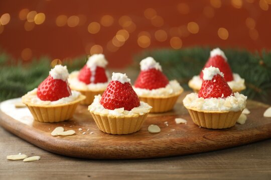 Christmas food. Tasty tartlets with strawberries, whipped cream, coconut flakes and decor on wooden table against background with blurred lights, closeup. Bokeh effect