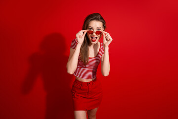 Obraz premium Young stylish woman in red outfit with sunglasses posing on a red background expressing surprise and joy