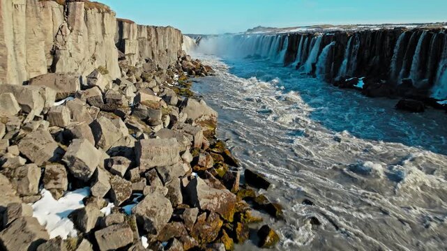 Aerial footage of Dettifoss waterfall in North Iceland. Water cascades forcefully through the basalt canyon surrounded by snow patches.