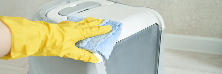 Gloved hand cleaning white trash can with blue cloth in home setting.