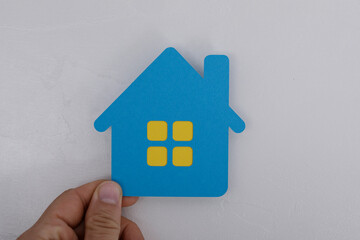 Close-up of hand holding a blue paper house cutout against a white, textured backdrop. Represents home, real estate, and concepts of ownership a