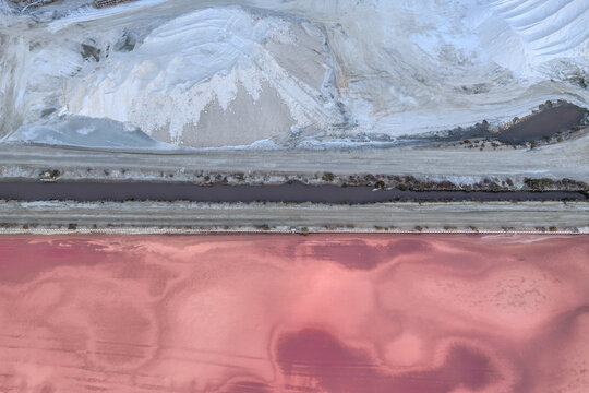 Aerial view of pink-hued waters reflecting the sky, contrasting sharply with the stark white salt piles and dark dividing line, Aigues, Occitanie, France.