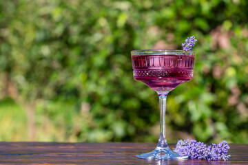 Bright cocktail or lemonade served in elegant crystal glass on wooden table against of a green summer garden. Purple color drink with lavender herb flowers, closeup