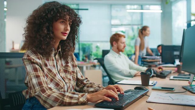 Pretty Caucasian woman in checkered shirt working at computer and looking thoughtfully at screen. Focused office worker concentrating on task while colleagues collaborating in bright modern workspace.