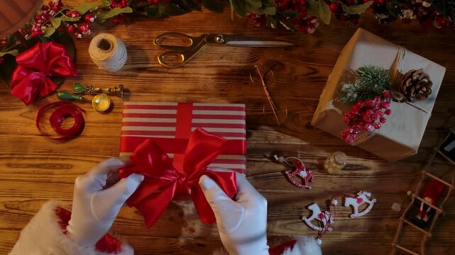Top view of Santa Claus's gloved hands creating festive gifts with his own hands on a rustic wooden table. Preparing for New Year's and Christmas celebrations.