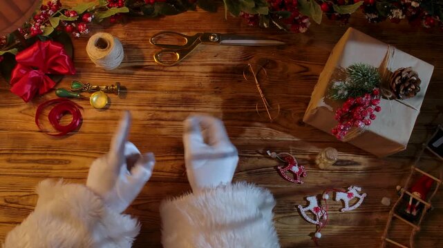 Top view of Santa Claus's gloved hands dancing merrily on a rustic wooden table. Preparations for New Year's and Christmas celebrations.